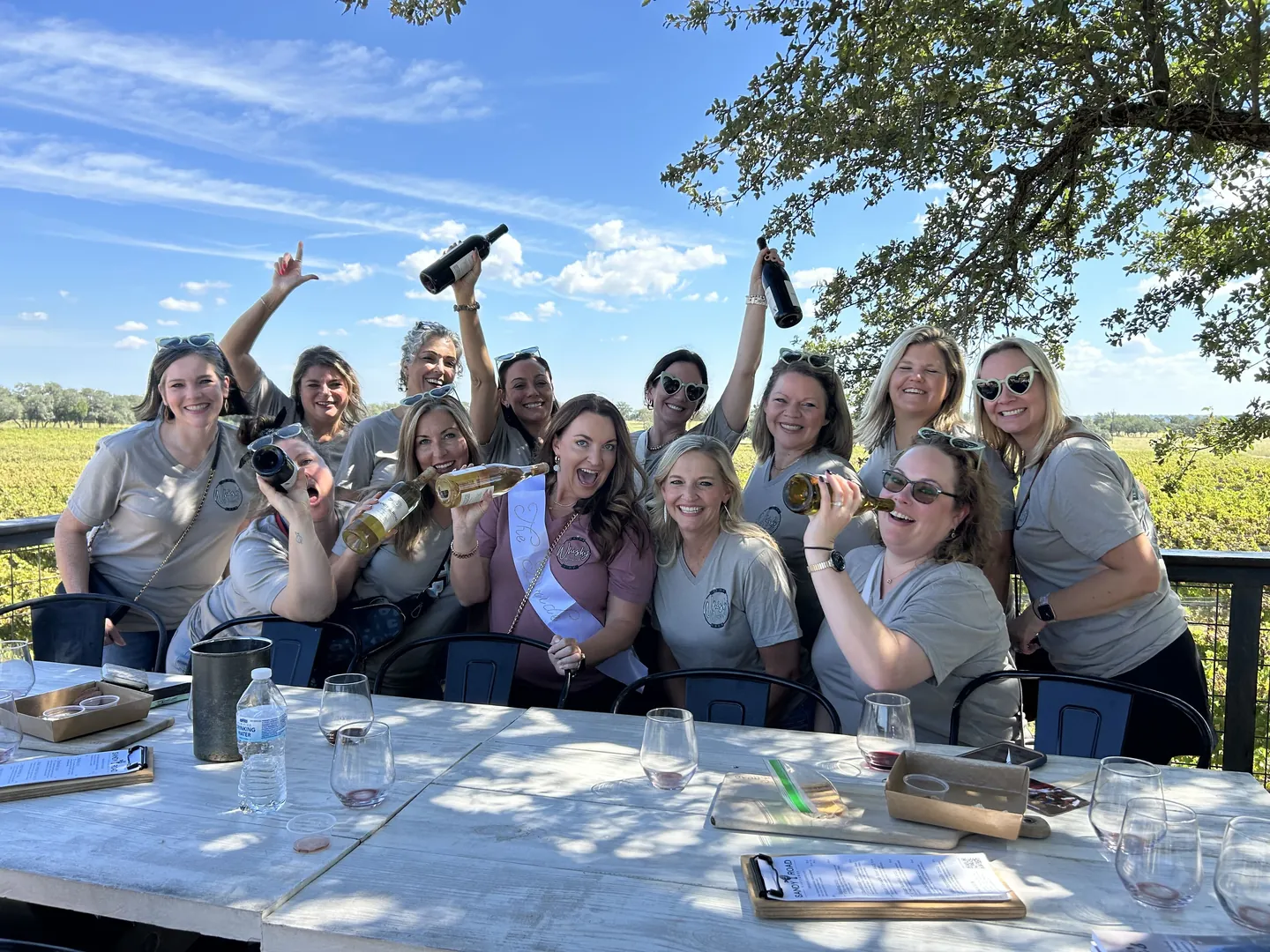 A bachelorette party on a winery deck under the oaks, bottles raised, the bride in her sash front and centre.